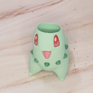 May include: A light green Bulbasaur-shaped planter. The planter has a round opening at the top and features the character's face with red eyes, a pink tongue, and green spikes. The planter is made of a smooth material.