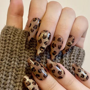 Könnte beinhalten: Eine Hand mit langen, mandelförmigen Nägeln. Die Nägel sind mit einer braunen Basis und einem Leopardenmuster in Dunkelbraun und Goldglitter bemalt. Ein modisches Accessoire.