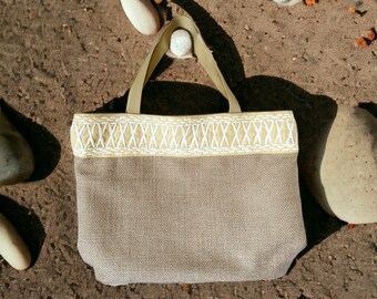 May include: A beige tote bag with a white embroidered trim. The bag is made of a woven fabric and has a wide, flat base.