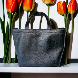 May include: A black ostrich leather tote bag with pink stitching. The bag has a top handle and a spacious interior.