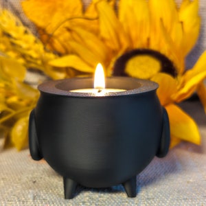 A Cauldron Tea light Holders in black, one featuring a real flame, set against an autumn-themed background adorned with sunflowers and pumpkins.