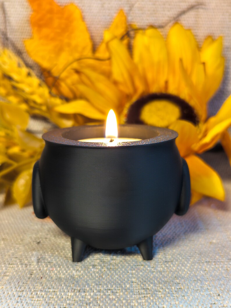 A Cauldron Tea light Holders in black, one featuring a real flame, set against an autumn-themed background adorned with sunflowers and pumpkins.