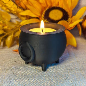 A Cauldron Tea light Holders in black, one featuring a real flame, set against an autumn-themed background adorned with sunflowers and pumpkins.