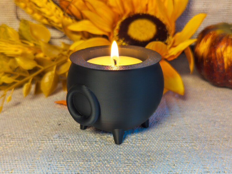 A Cauldron Tea light Holders in black, one featuring a real flame, set against an autumn-themed background adorned with sunflowers and pumpkins.