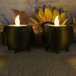 Two black Cauldron Tea light Holders with lit candles are placed on a textured surface, surrounded by autumn-themed decor featuring a sunflower and dried foliage.