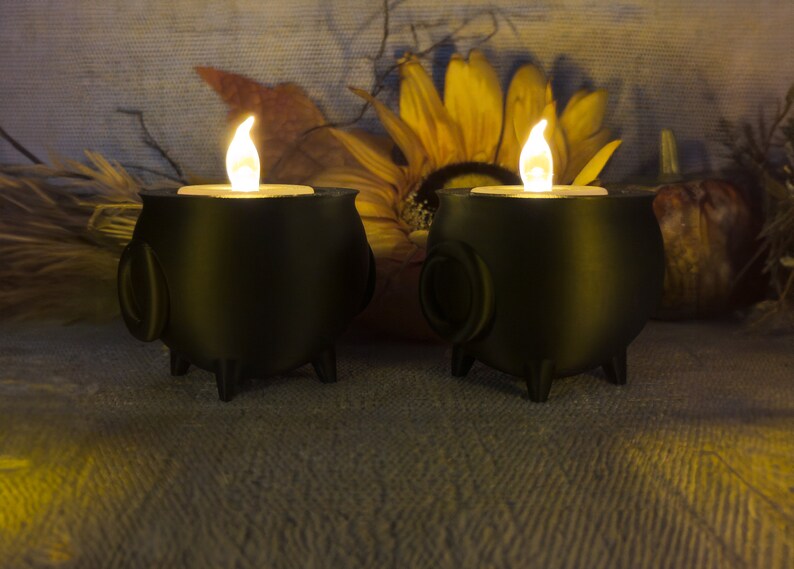 Two black Cauldron Tea light Holders with lit candles are placed on a textured surface, surrounded by autumn-themed decor featuring a sunflower and dried foliage.