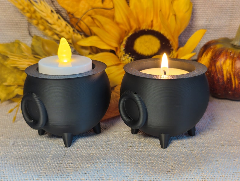 Two Cauldron Tea light Holders in black, one featuring a plastic flame and the other with a real flame, set against an autumn-themed background adorned with sunflowers and pumpkins.