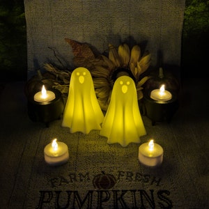 Two White 3D Printed glowing ghost decorations are surrounded by four lit candles, placed on a textured cloth that reads "Farm Fresh Pumpkins" & with a sunflower and small pumpkins in the background.