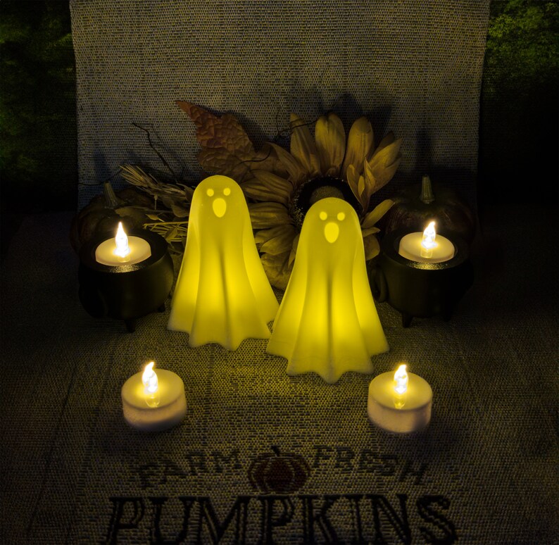 Two White 3D Printed glowing ghost decorations are surrounded by four lit candles, placed on a textured cloth that reads "Farm Fresh Pumpkins" & with a sunflower and small pumpkins in the background.