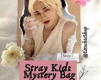 May include: A promotional image featuring a light-haired person in a cream-colored top, set against a torn paper backdrop. The text "Stray Kids Mystery Bag" is displayed on a pink banner. Decorative flowers and the text "@LitschisShop" are also visible.