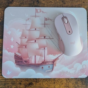 May include: A pink mouse pad with a pink and white ship sailing on clouds. The text "To Wonderland..." is printed on the bottom right corner. A pink wireless mouse with the Logitech logo is on the mouse pad.