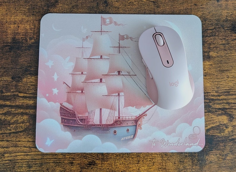 May include: A pink mouse pad with a pink and white ship sailing on clouds. The text "To Wonderland..." is printed on the bottom right corner. A pink wireless mouse with the Logitech logo is on the mouse pad.