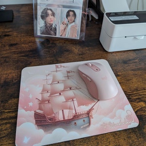 May include: Pink mousepad with a pink mouse on top. The mousepad features a pink and white illustration of a ship sailing on clouds with the text "To Wonderland..." and "ATEEZ" on it.