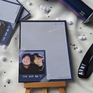 Può includere: Un blocco note con un design a stelle blu e bianco, con una foto di due persone. Il blocco note ha il testo "I GOT YOU" e "YOU GOT ME". Il blocco note è esposto su un piccolo cavalletto di legno.