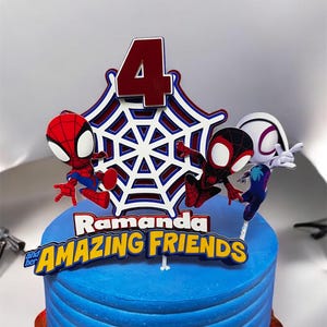 May include: A blue and white cake with a Spiderman theme. The cake is decorated with a red number 4, a white spiderweb, and three Spiderman figures. The cake is also decorated with the text "Ramanda and her AMAZING FRIENDS".