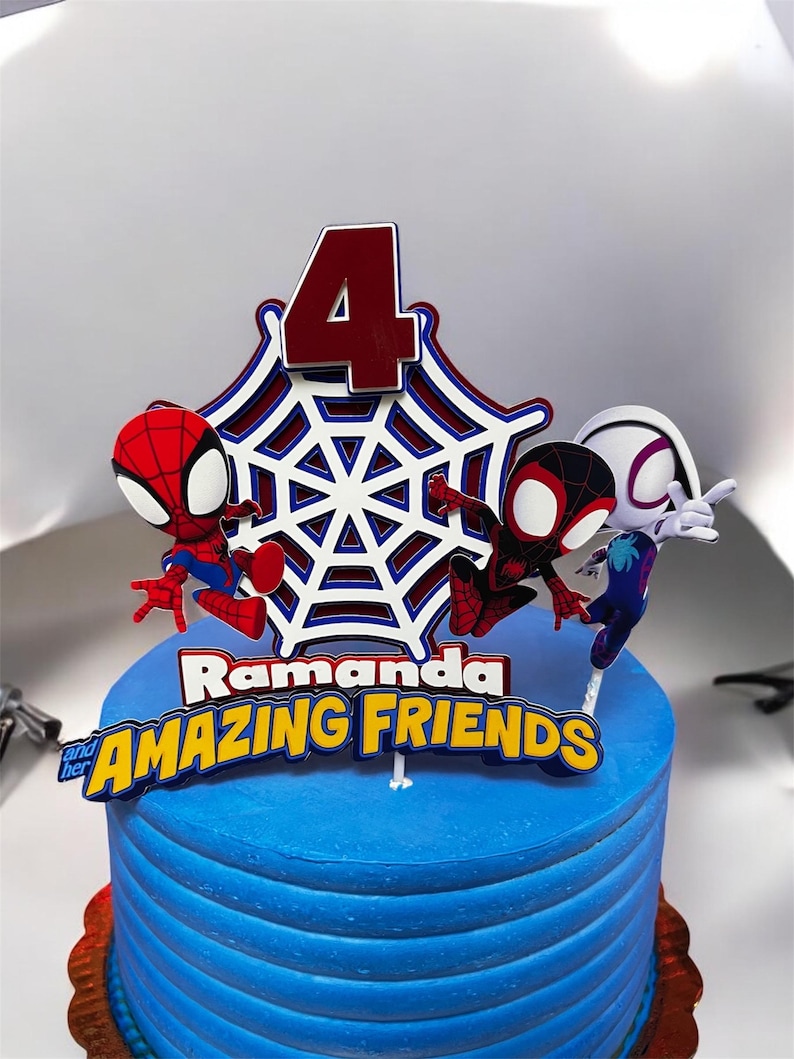 May include: A blue and white cake with a Spiderman theme. The cake is decorated with a red number 4, a white spiderweb, and three Spiderman figures. The cake is also decorated with the text "Ramanda and her AMAZING FRIENDS".