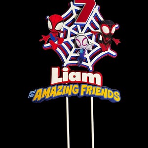 May include: A red, white and blue cake topper with a spiderweb design. Three Spiderman characters are on the web, and the number 7 is in the centre. The text "Liam and his AMAZING FRIENDS" is below the web.