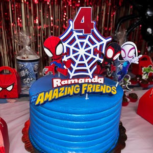May include: A blue and white frosted cake with a Spiderman theme. The cake is decorated with a web, a number 4, and three Spiderman characters. The cake is on a white cake stand with a scalloped edge. The cake is decorated with the words "Ramanda and her AMAZING FRIENDS".
