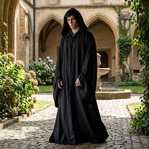 Peut inclure: Une cape noire à capuche avec de longues manches. La cape est faite d'un tissu fluide et est portée par une personne debout sur un chemin pavé. L'arrière-plan présente un bâtiment voûté et de la verdure.