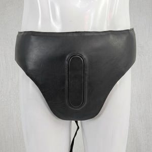 May include: A black leather protective groin guard featuring a smooth, matte finish and a central, vertically oriented padded oval section. The piece is constructed with reinforced stitching around the edges and a single thin strap extending downward.
