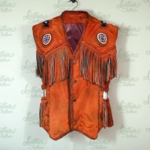 May include: An orange suede vest with fringe detailing and decorative patches. The vest has a button-up front and is hanging on a black hanger. The vest is a traditional style.