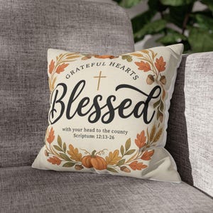 May include: A square throw pillow with a cream-colored cover featuring a central black script text that reads Blessed, a small cross, and the text Grateful Hearts with your head to the county Scripture 12:13-26, surrounded by autumn leaves and pumpkins.