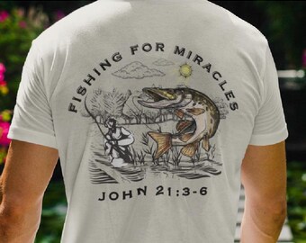 Peut inclure: T-shirt blanc avec une illustration en noir et blanc d'un pêcheur et d'un poisson sautant hors de l'eau. Le texte "Fishing for Miracles" est écrit en cercle autour de l'illustration. Le texte "John 21:3-6" est écrit sous l'illustration.