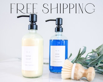 May include: Two glass pump bottles with black pumps, one filled with a clear yellow liquid and labeled "HAND SOAP" and the other filled with a blue liquid and labeled "DISH SOAP". A wooden scrub brush with white bristles is in the foreground.