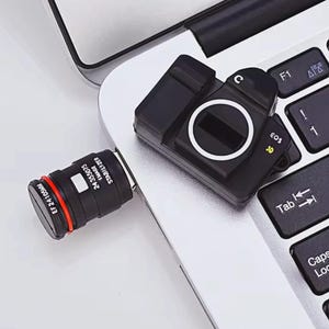 May include: A black USB flash drive in the shape of a camera lens and a camera body, connected to a laptop. The lens is marked with "EF 24 105MM" and "24 355075 IMAGE STABILIZER". The camera body has the text "EOS 30".