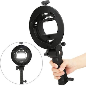 Puede incluir: Un adaptador de flash anular negro para fotografía, sostenido por una mano. El adaptador tiene un diseño circular con una abertura central y un asa. Incluye perillas de ajuste y un mecanismo de montaje.