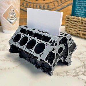 Peut inclure: Un porte-cartes de visite en forme de bloc moteur imprimé en 3D gris et noir. Le porte-cartes a une fente pour les cartes de visite et est conçu pour ressembler à un moteur de voiture.