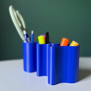 May include: Three cobalt blue desk organizers with a wavy design. The organizers hold pens, scissors, and markers in various colors. The containers are cylindrical with a unique, curved top edge. The background is a dark green.