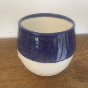 May include: A small, rounded ceramic cup featuring a two-tone design. The upper portion is finished in a deep, speckled blue glaze, while the lower section remains a smooth, matte off-white. The interior of the cup is a plain, light cream colour.