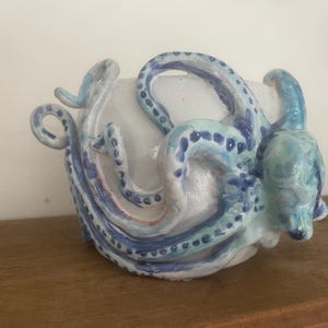 Op de afbeelding: Een witte keramische kom met een gebeeldhouwde octopus aan de buitenkant. De octopus is beschilderd in tinten blauw en lichtblauw met zichtbare zuignapdetails langs de tentakels die rond het oppervlak van de kom krullen.