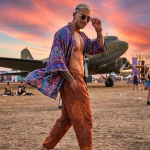 May include: A loose-fitting, open-front kimono with a vibrant blue, purple, and red patterned design, paired with rust-colored joggers featuring a circular geometric print, worn with simple tan flip-flops and round-framed sunglasses.
