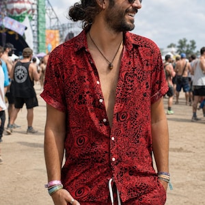 May include: Red button-down shirt and matching shorts set with a black eye and floral pattern. The short-sleeved shirt is unbuttoned, and the shorts have a drawstring. The outfit is worn at an outdoor event.