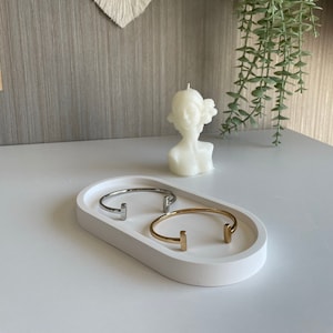 May include: A white oval tray holds two metal bracelets, one silver and one gold, alongside a white bust candle. A macrame wall hanging and hanging greenery are in the background. The bracelets have a T-shaped design.