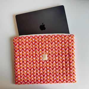 Può includere: Un laptop nero con il logo Apple è all'interno di una custodia per laptop trapuntata aperta. La custodia presenta un motivo a onde ripetute nei toni dell'arancione e del rosa. Una piccola etichetta rettangolare è cucita sul davanti.
