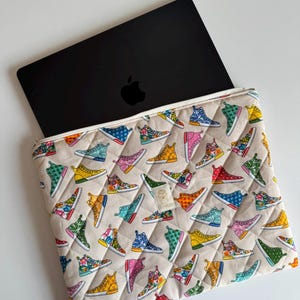 Può includere: Un computer portatile nero parzialmente inserito in una custodia imbottita beige. La custodia è decorata con sneakers colorate in vari design. La custodia ha un bordo bianco e una piccola etichetta con testo.