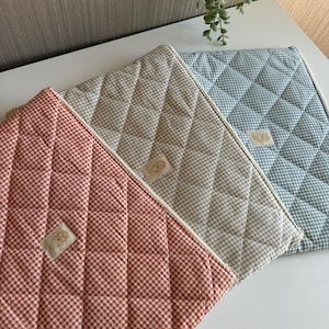 Puede incluir: Tres manteles individuales acolchados con estampado de cuadros. Los manteles individuales son de color rojo, beige y azul. Cada mantel individual tiene un patrón de costura de diamante y una pequeña etiqueta. Sobre una superficie blanca.