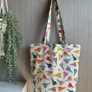 May include: A beige tote bag with a repeating pattern of colorful sneakers. The sneakers are in various colors including red, blue, green, yellow, and pink. The bag has matching handles and a small tag.