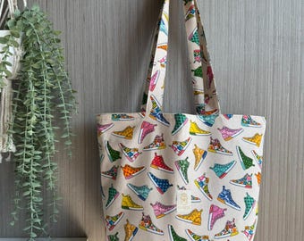 Può includere: Una borsa tote beige con un motivo ripetuto di sneakers colorate. Le sneakers sono in vari colori tra cui rosso, blu, verde, giallo e rosa. La borsa ha manici abbinati.