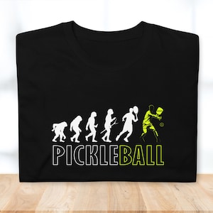 May include: Black t-shirt with a white silhouette of the evolution of humans ending with a person playing pickleball. The text "PICKLEBALL" is written in white below the image.