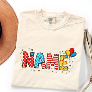 May include: A cream-colored t-shirt with a Comfort Colors label featuring the word NAME in large, colorful, patterned block letters. The letters have a stitched-look border and are decorated with red polka dots, blue dots, red stripes, and yellow confetti patterns, accompanied by three balloons.