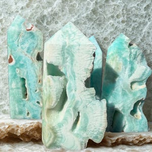May include: Three turquoise-colored aragonite crystal towers with unique patterns and textures. The crystals have a mix of light and dark blue hues, with some areas appearing almost white. The crystals are displayed on a light-colored, textured surface.