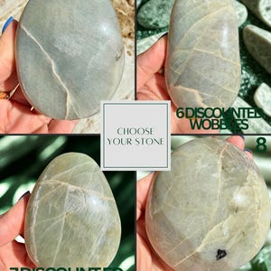 May include: Four smooth, oval-shaped stones in shades of gray and green are displayed. Each stone has unique veining patterns. The image includes text that reads "Choose Your Stone" and numbers 5, 6, 7, and 8, along with the words "Discounted Wobbles".