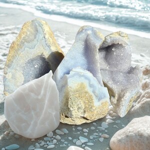 May include: A selection of natural crystals and geodes in white, blue, and gold hues, displayed on a sandy surface with small pebbles. The background shows the ocean with gentle waves.