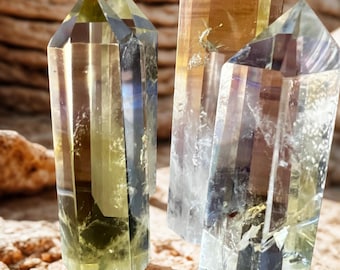 May include: Four clear quartz crystal points with yellow and brown hues. The crystals are faceted and stand upright on a textured, light brown surface. The crystals have a natural, earthy appearance.