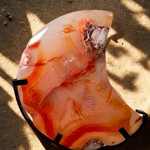 May include: A polished, translucent agate stone with swirling patterns of orange, red, and white, mounted on a black metal stand. The stone's unique shape and color variations are highlighted by the natural light.