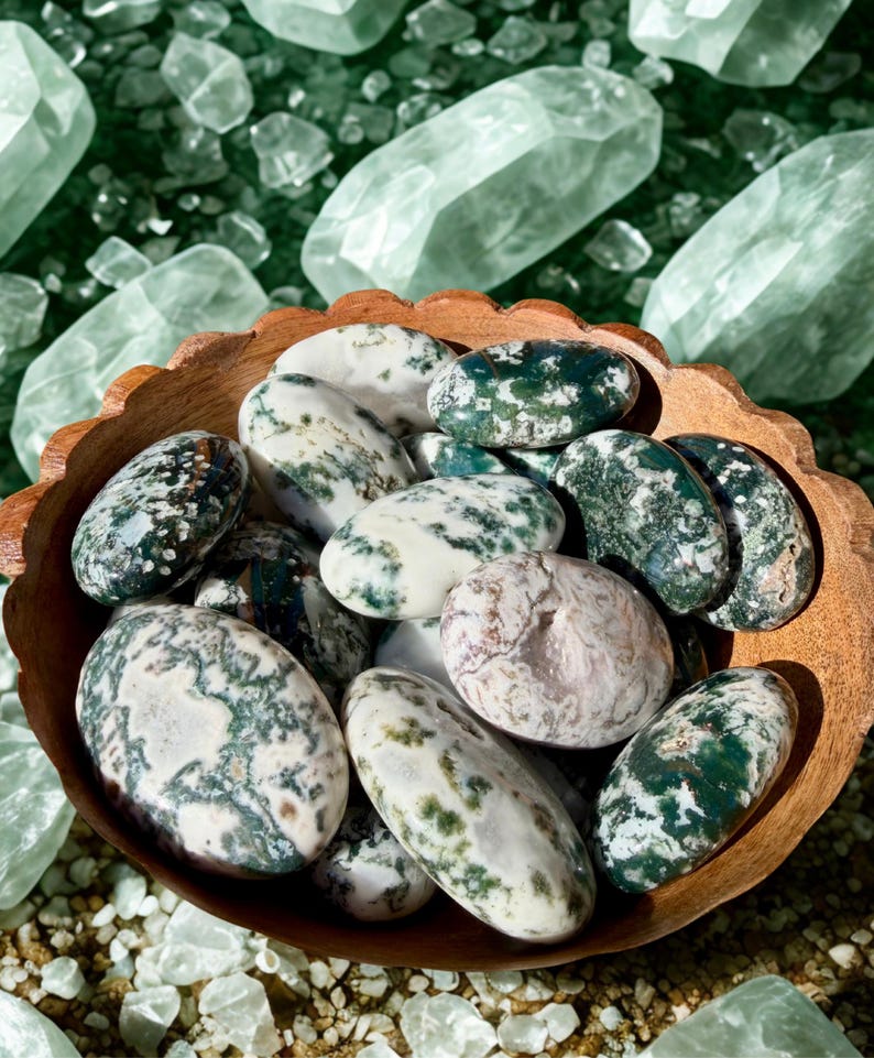 May include: A wooden bowl filled with polished moss agate stones. The stones are oval-shaped with a mix of white, green, and brown patterns. Large, clear quartz crystals are scattered in the background, creating a natural, earthy aesthetic.